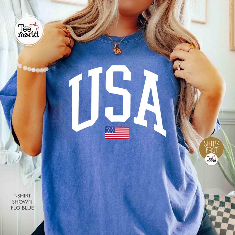 May include: Blue t-shirt with the word "USA" in large white block letters and a small American flag graphic. The shirt is a solid blue color and has a casual, relaxed fit.