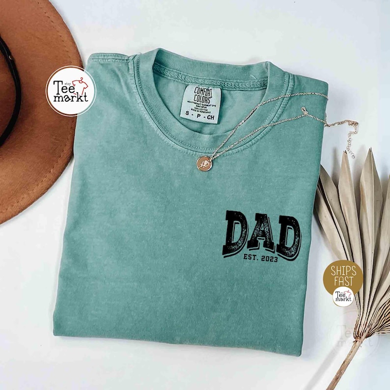 May include: A seafoam green t-shirt with the word "DAD" in black block letters and "EST. 2023" below. A silver necklace with a pendant rests on the shirt. The shirt has a tag that says "COMFORT COLORS".