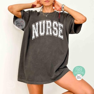 May include: Dark gray t-shirt with the word "NURSE" in large, white, block letters across the chest. The shirt has a vintage, slightly faded look. The tee is a relaxed fit.
