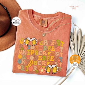 K&ouml;nnte beinhalten: Ein korallenfarbenes T-Shirt mit dem Wort "OKTOBERFEST" in einem Retro-Schriftzug, verziert mit Bierkr&uuml;gen und Blumen. Das Shirt hat auch den Text "PERFEST" und "OKTOBERFEST" in einem sich wiederholenden Muster. Das Shirt hat einen runden Ausschnitt und kurze &Auml;rmel.