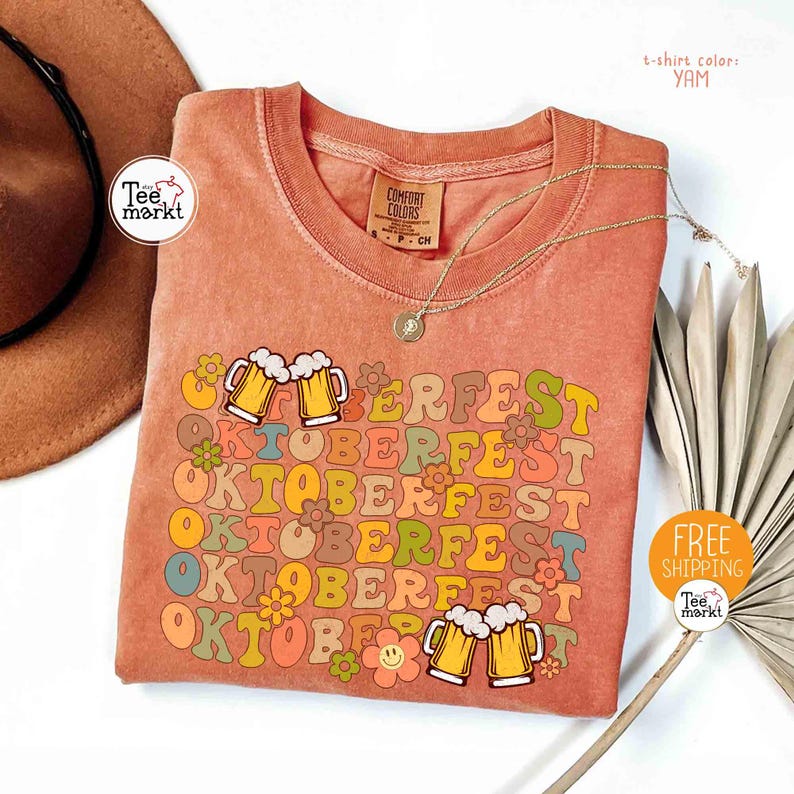 K&ouml;nnte beinhalten: Ein korallenfarbenes T-Shirt mit dem Wort "OKTOBERFEST" in einem Retro-Schriftzug, verziert mit Bierkr&uuml;gen und Blumen. Das Shirt hat auch den Text "PERFEST" und "OKTOBERFEST" in einem sich wiederholenden Muster. Das Shirt hat einen runden Ausschnitt und kurze &Auml;rmel.