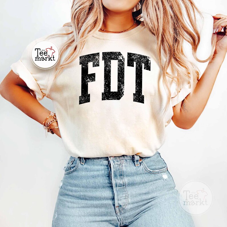 May include: Cream-colored t-shirt with the letters "FDT" in a distressed black font. The shirt has short sleeves and is paired with blue jeans. A small logo is visible on the left sleeve.