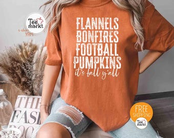 May include: An orange t-shirt with the words "FLANNELS BONFIRES FOOTBALL PUMPKINS it's fall y'all" in white. The shirt has short sleeves and is worn with ripped blue jeans. A logo is visible in the upper left corner.