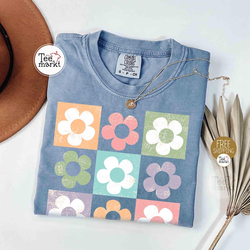 May include: A blue Comfort Colors t-shirt featuring a grid of colorful flowers in orange, pink, green, white, blue, and purple squares. A gold necklace and a brown hat are also visible.