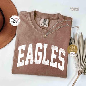 May include: Espresso-colored t-shirt with the word "EAGLES" in distressed white lettering. The shirt has a crew neck and short sleeves. A silver necklace with a pendant is draped on the shirt. The shirt is part of the "Tee markt" collection.