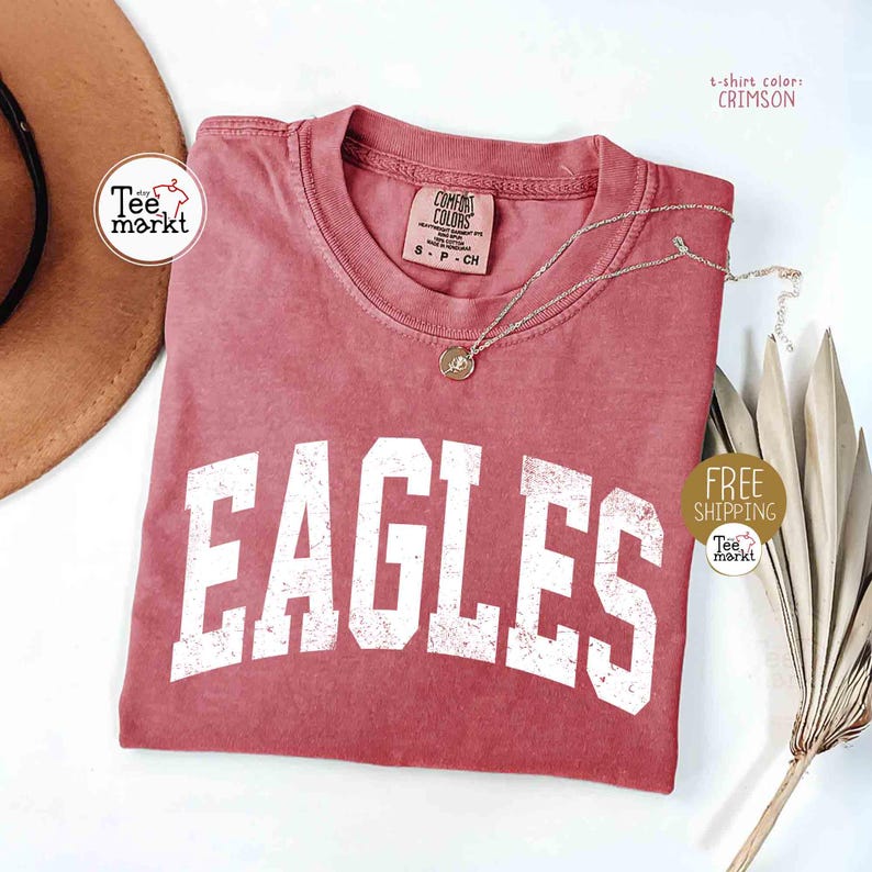 May include: Crimson-colored t-shirt with the word "EAGLES" in large, distressed white letters. A gold pendant necklace rests on the shirt. The shirt has a tag that says "COMFORT COLORS".