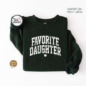 May include: A forest green crewneck sweatshirt featuring the white distressed text FAVORITE DAUGHTER in an arched collegiate font with a small white heart icon centred below the lettering. The garment has long sleeves and a ribbed neckline.