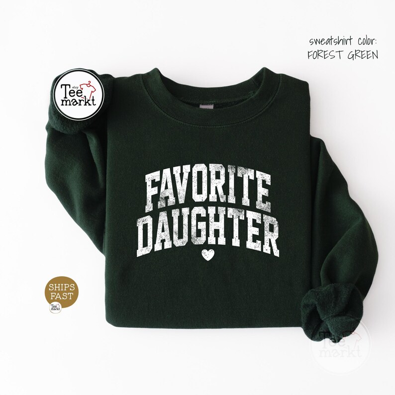 May include: A forest green crewneck sweatshirt featuring the white distressed text FAVORITE DAUGHTER in an arched collegiate font with a small white heart icon centred below the lettering. The garment has long sleeves and a ribbed neckline.