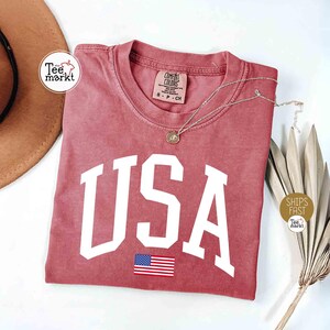 May include: A dusty rose-colored t-shirt with the word "USA" in large white block letters and a small American flag graphic. A silver necklace with a pendant rests on the shirt. The shirt is folded on a white surface.