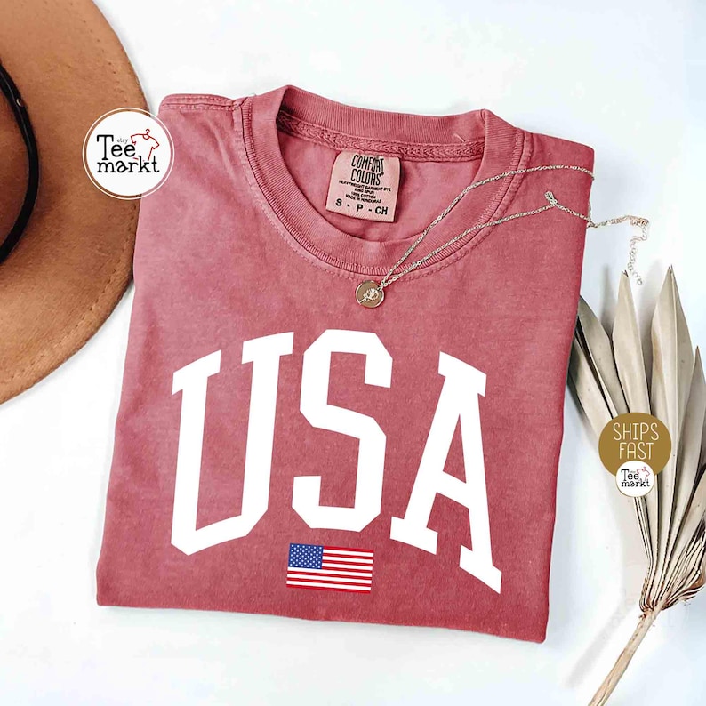 May include: A dusty rose-colored t-shirt with the word "USA" in large white block letters and a small American flag graphic. A silver necklace with a pendant rests on the shirt. The shirt is folded on a white surface.