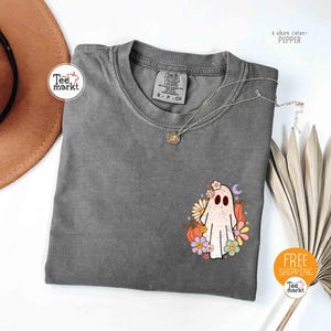 以下が含まれることがあります： ペッパーカラーのTシャツ。花のゴーストグラフィックが特徴です。デザインは、花の冠をかぶった白いゴースト、カラフルな花、カボチャ、三日月が特徴です。ペンダント付きのシルバーネックレスがシャツの上にあります。