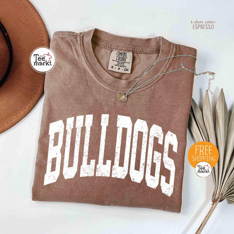 May include: Espresso-colored t-shirt with the word "BULLDOGS" in white block letters. The shirt has a crew neck and short sleeves. A silver necklace with a pendant rests on the shirt. The shirt is displayed with a brown hat and dried leaves.