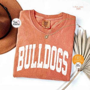 May include: A yam-colored t-shirt with the word "BULLDOGS" in large white block letters. A gold necklace with a pendant rests on the shirt. A brown hat and dried plant are in the background.