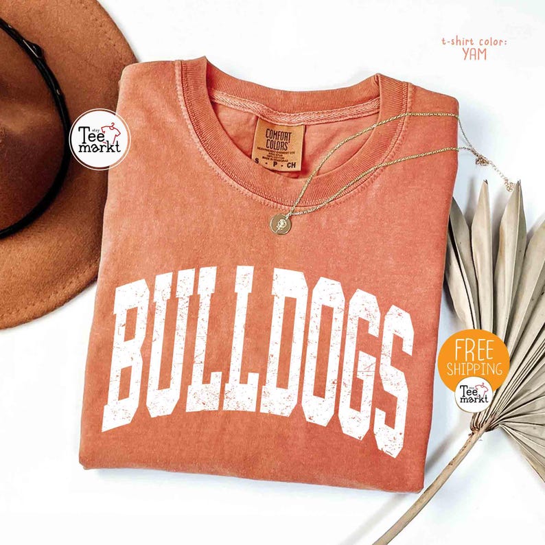 May include: A yam-colored t-shirt with the word "BULLDOGS" in large white block letters. A gold necklace with a pendant rests on the shirt. A brown hat and dried plant are in the background.