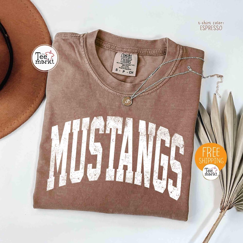 May include: Espresso-colored t-shirt with the word "MUSTANGS" in distressed white lettering. A silver necklace with a pendant rests on the shirt. The shirt is a Comfort Colors brand, size S-P-CH.