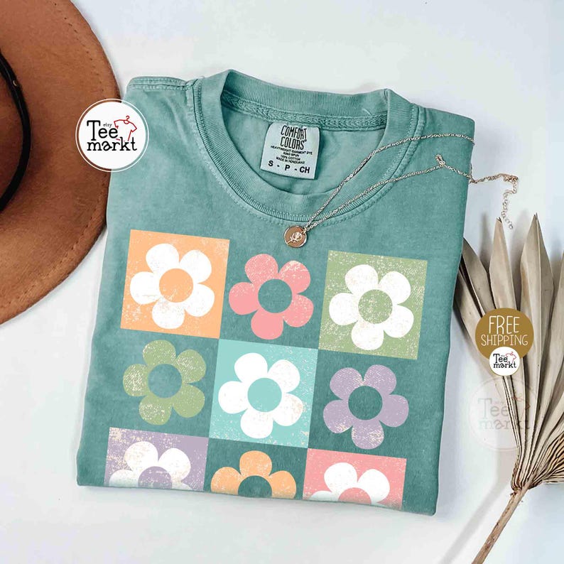 May include: A teal blue t-shirt with a graphic print of nine pastel colored flowers in a grid pattern. The t-shirt has a tag that reads 'Comfort Colors' and 'S-P-CH'.