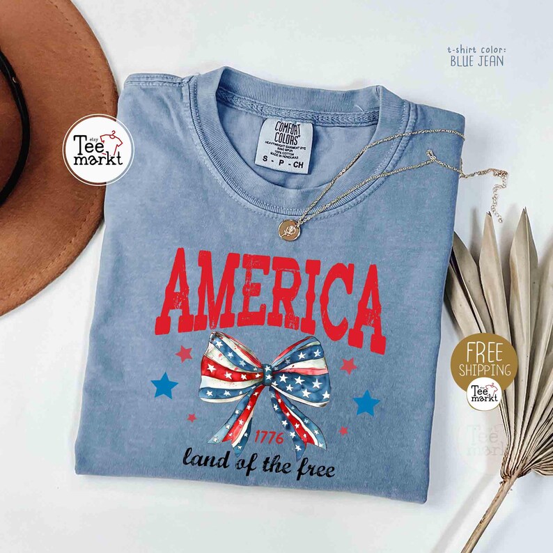 May include: Blue jean t-shirt with the word "AMERICA" in red, a bow with a stars and stripes design, and the text "1776 land of the free." A gold necklace and a brown hat are also visible.