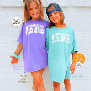 May include: Two young people wearing oversized t-shirts with the word "MUSTANGS" in white block letters. One shirt is violet, the other is chalky mint. The image includes the text "YOUTH SIZES AVAILABLE".
