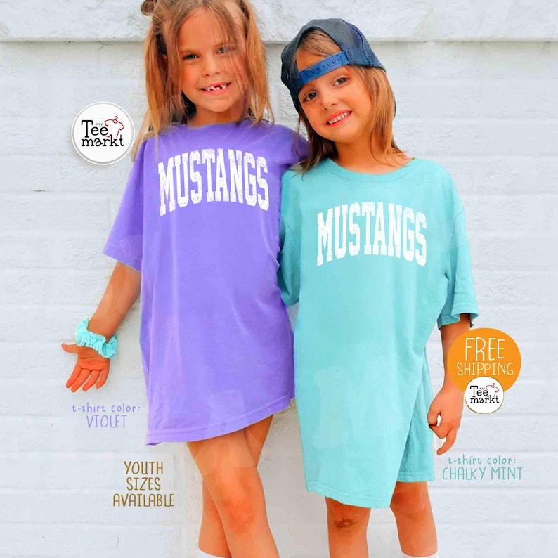 May include: Two young people wearing oversized t-shirts with the word "MUSTANGS" in white block letters. One shirt is violet, the other is chalky mint. The image includes the text "YOUTH SIZES AVAILABLE".