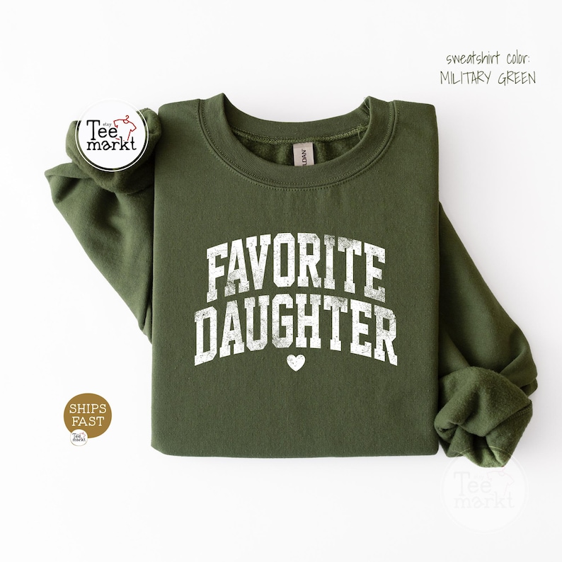 May include: A military green long-sleeve sweatshirt featuring the white distressed text "FAVORITE DAUGHTER" arched across the chest, accompanied by a small white heart icon centred below the lettering. The garment has a crew neckline.