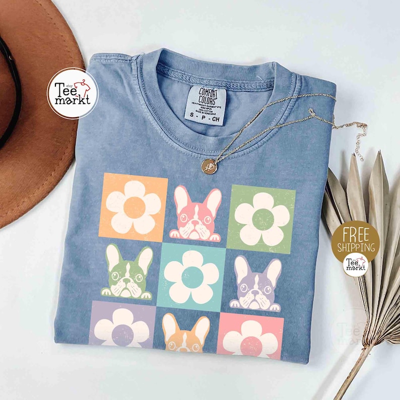 May include: A light blue t-shirt featuring a colorful design of flowers and French bulldogs. The shirt has a crew neck and short sleeves. A gold necklace with a pendant is draped across the shirt. The shirt has the text "Comfort Colors" on the tag.