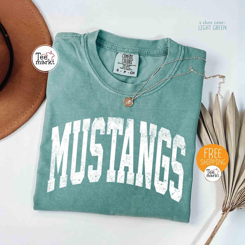 May include: Light green t-shirt with the word "MUSTANGS" in large, distressed white letters. The shirt has a crew neck and is folded. A silver necklace with a pendant is on the shirt. The shirt is from Comfort Colors.