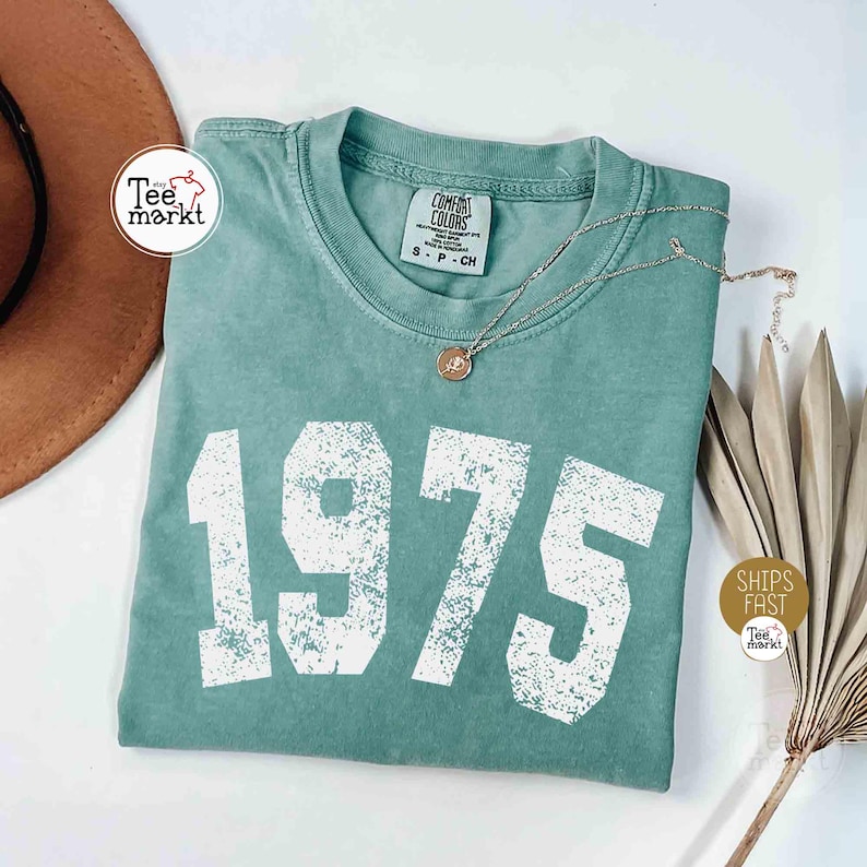 May include: A seafoam green t-shirt with the number "1975" printed in white. The shirt has a crew neck and short sleeves. A silver necklace with a small pendant rests on the shirt. A brown hat and dried plant are in the background.