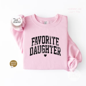 May include: A light pink crewneck sweatshirt featuring the text FAVORITE DAUGHTER in a distressed black font with a small black heart icon centred below the lettering. The garment has long sleeves and a ribbed collar, cuffs, and hem.