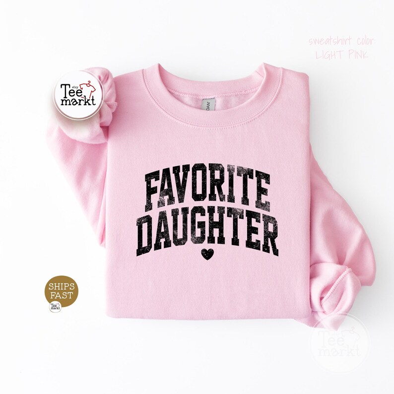 May include: A light pink crewneck sweatshirt featuring the text FAVORITE DAUGHTER in a distressed black font with a small black heart icon centred below the lettering. The garment has long sleeves and a ribbed collar, cuffs, and hem.
