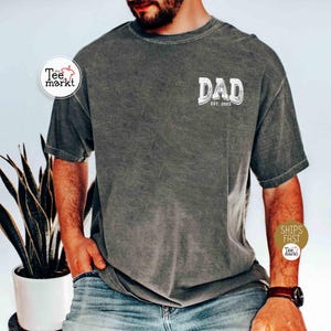 May include: Dark gray t-shirt with the word "DAD" in a distressed white font, with "EST. 2023" below it. The shirt is a solid color and has short sleeves. The shirt is made of a soft, comfortable material.