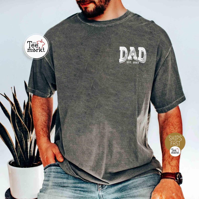 May include: Dark gray t-shirt with the word "DAD" in a distressed white font, with "EST. 2023" below it. The shirt is a solid color and has short sleeves. The shirt is made of a soft, comfortable material.