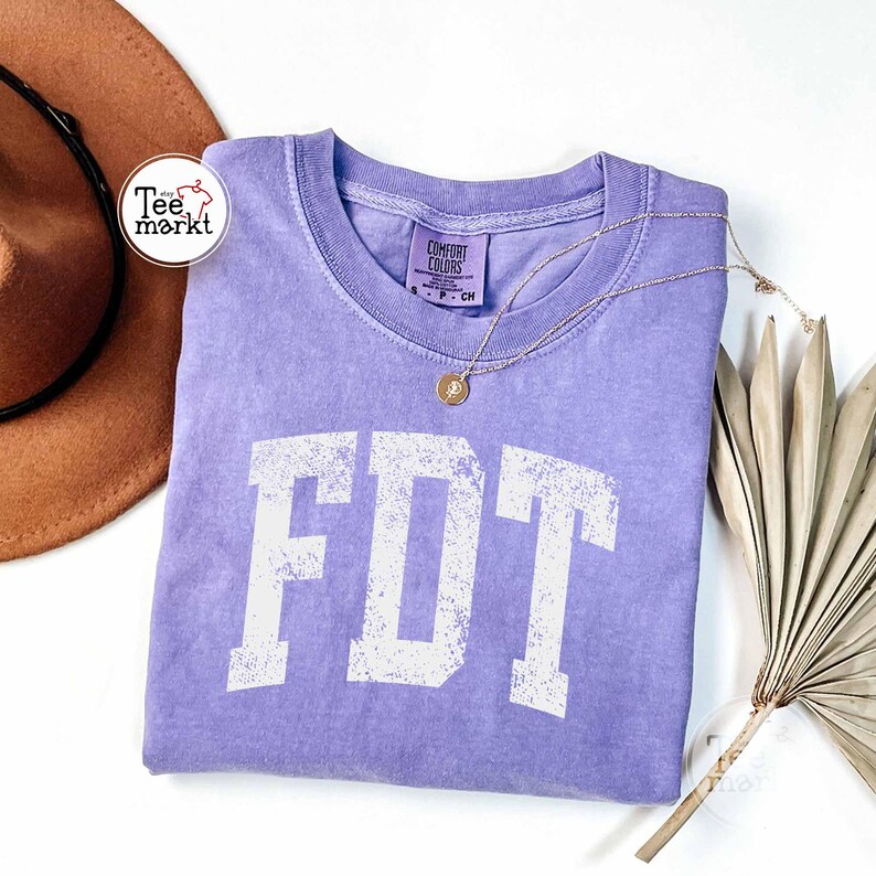 May include: A lavender Comfort Colors t-shirt with the white letters "FDT" printed on the front. A gold necklace with a pendant rests on the shirt. A brown hat and dried plant are in the background.