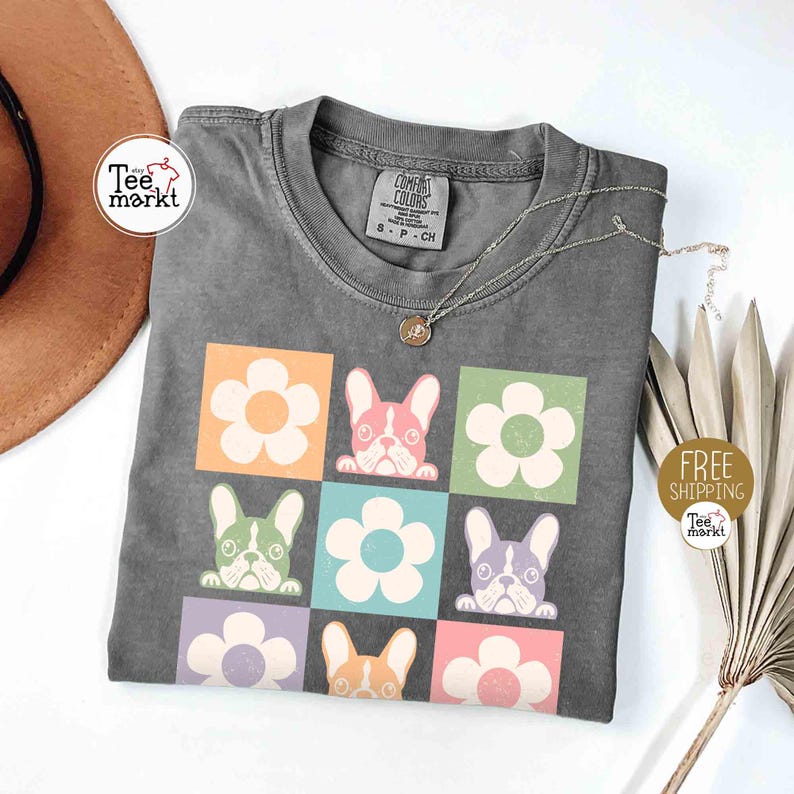 May include: Gray t-shirt featuring a colorful graphic of French bulldogs and flowers. The design includes a grid of squares with different colored backgrounds, each containing a flower or a dog. A silver necklace with a pendant is on the shirt.