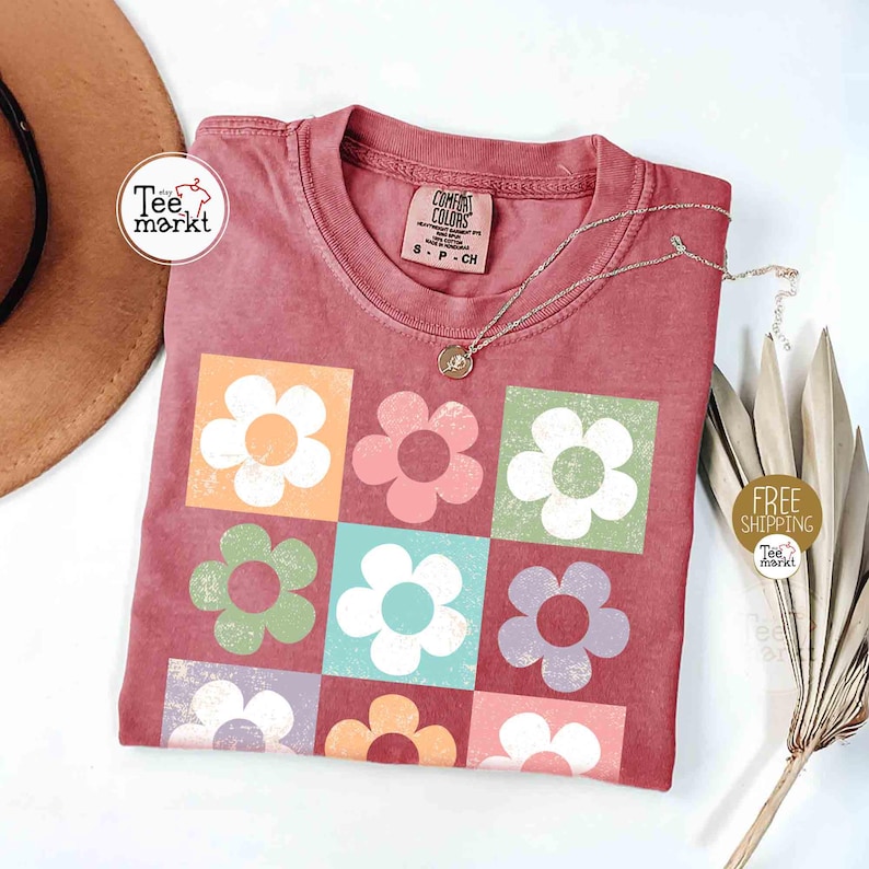 May include: A dusty rose-colored t-shirt with a square floral pattern in various colors. The shirt has a crew neck and short sleeves. A silver necklace with a pendant is draped on the shirt. The tag reads "Comfort Colors".