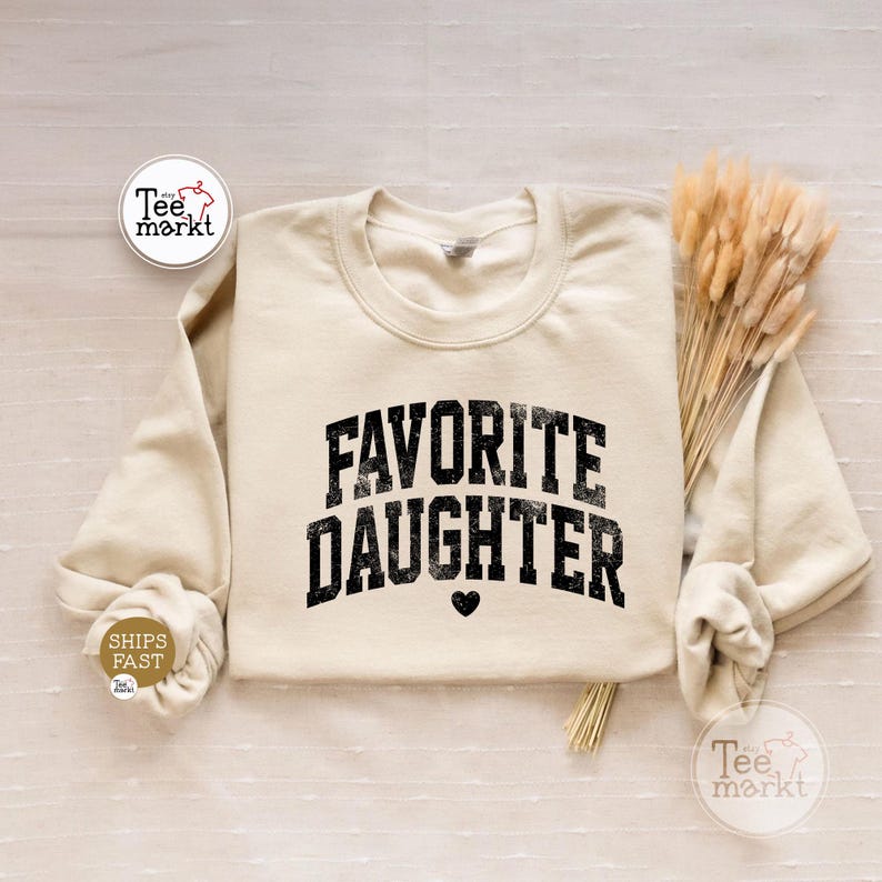 May include: A cream-coloured crewneck sweatshirt featuring the black text FAVORITE DAUGHTER in a distressed, arched varsity-style font with a small black heart icon centred below the lettering. The garment has long sleeves and a ribbed neckline.