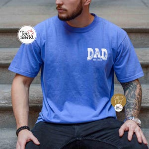 May include: A blue t-shirt with the word "DAD" in white, with "EST. 2023" below it. The shirt is short-sleeved and has a relaxed fit. The person is wearing black pants and a bracelet.