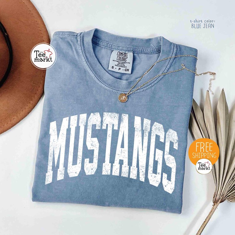 May include: A blue jean colored t-shirt with the word "MUSTANGS" in white block letters. The shirt has a crew neck and short sleeves. A gold necklace with a pendant is draped across the shirt. The shirt is folded on a white surface.