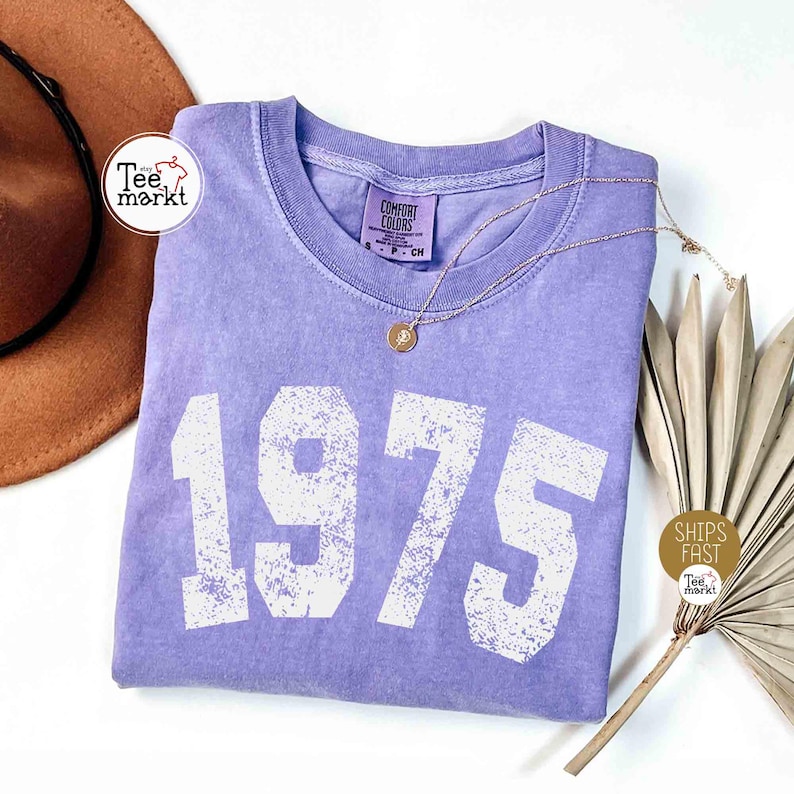 May include: A lavender-colored t-shirt with the number "1975" printed in white. The shirt has a crew neck and is adorned with a gold necklace. A brown hat and dried plant are in the background.