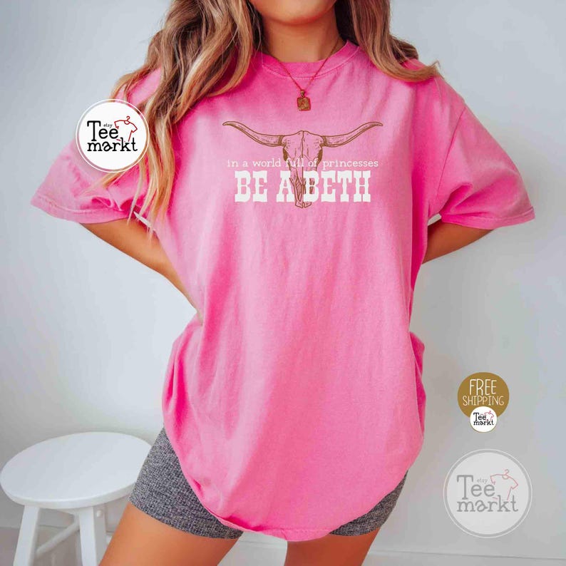 Puede incluir: Camiseta rosa brillante con el texto "in a world full of princesses BE A BETH" y un gr&aacute;fico de calavera de cuernos largos. La camiseta tiene mangas cortas y cuello redondo. El logo de Tee markt es visible.