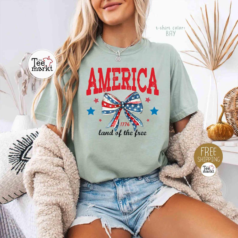 May include: A light green t-shirt with the word "AMERICA" in red block letters. Below the word is a bow in red, white, and blue, with the text "1776 land of the free". The shirt has a relaxed fit.