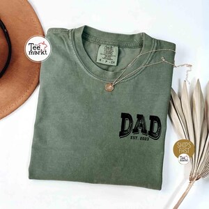 May include: An olive green t-shirt with the word "DAD" in black, with "EST. 2023" below it. A gold necklace with a pendant rests on the shirt. The shirt is folded on a white surface.