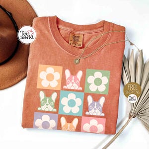 May include: Orange t-shirt with a square pattern of flowers and French bulldog faces in various colors. The shirt has a crew neck and short sleeves. A gold necklace with a small pendant is draped across the shirt. The words "Tee Markt" are printed on a white circle.
