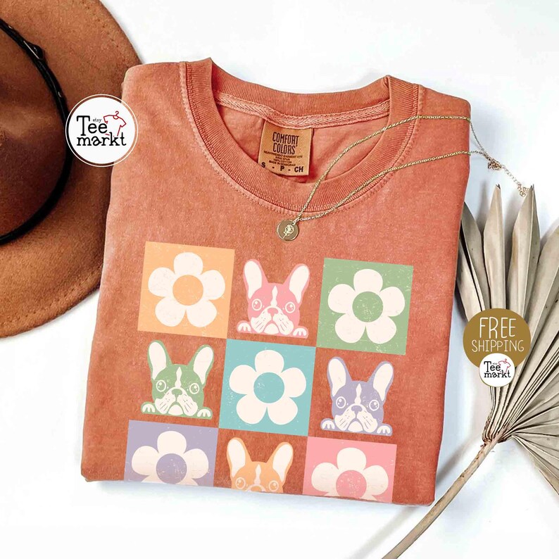 May include: Orange t-shirt with a square pattern of flowers and French bulldog faces in various colors. The shirt has a crew neck and short sleeves. A gold necklace with a small pendant is draped across the shirt. The words "Tee Markt" are printed on a white circle.