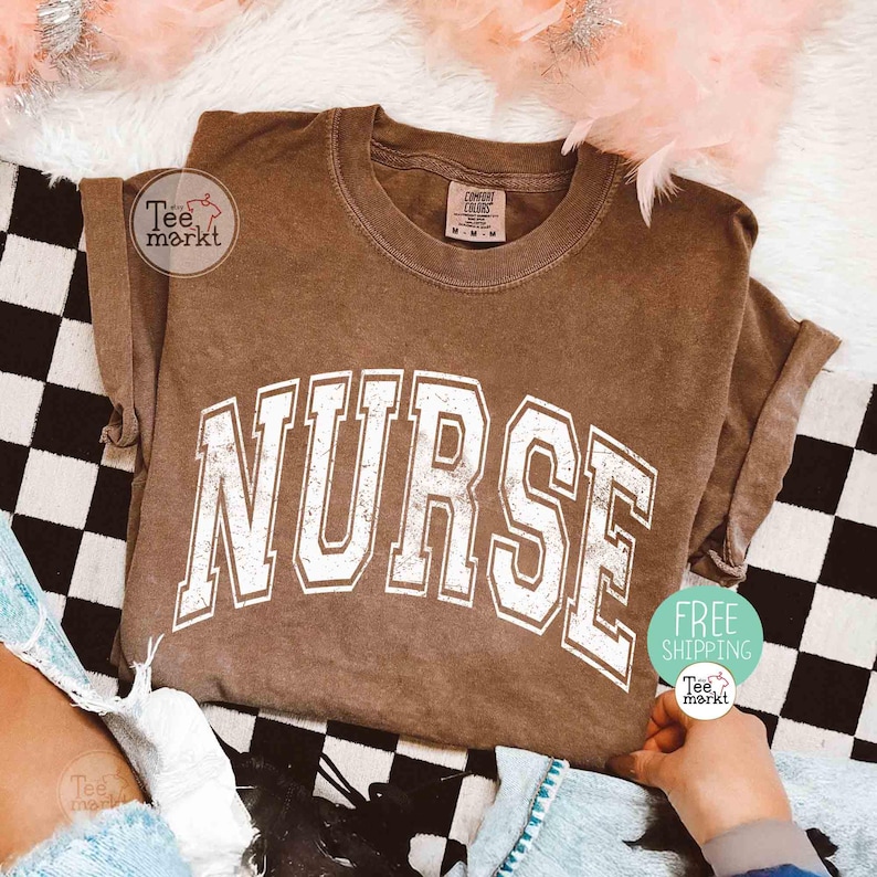 May include: A brown t-shirt with the word "NURSE" in large, white, collegiate-style lettering. The shirt is folded and rests on a black and white checkered surface. The shirt has a vintage, worn look.