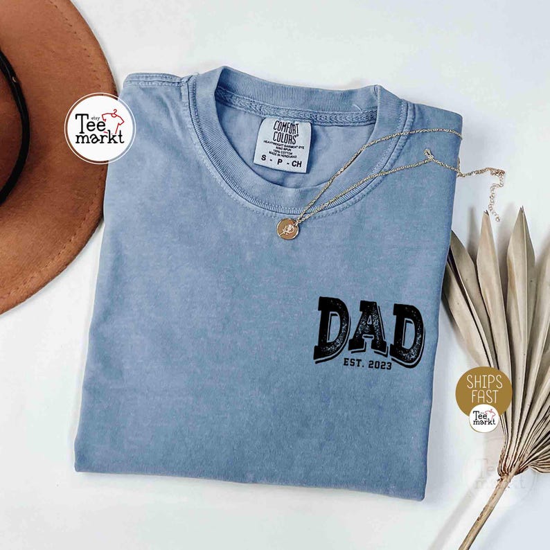 May include: A light blue t-shirt with the word "DAD" in black, and "EST. 2023" below it. A gold necklace with a pendant rests on the shirt. The shirt is on a white surface with a brown hat and dried plant stems.