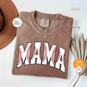 May include: Espresso-coloured t-shirt with the word "MAMA" in white, baseball-style lettering with red stitching. The shirt has a crew neck and short sleeves. A silver necklace with a pendant is draped across the shirt. The shirt is from Tee Markt.