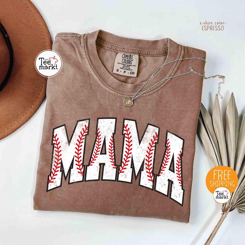 May include: Espresso-coloured t-shirt with the word "MAMA" in white, baseball-style lettering with red stitching. The shirt has a crew neck and short sleeves. A silver necklace with a pendant is draped across the shirt. The shirt is from Tee Markt.