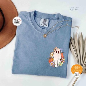 以下が含まれることがあります： 青いジーンズカラーのTシャツ。ゴーストのグラフィックが特徴です。ゴーストは花、カボチャ、三日月で飾られています。Tシャツは丸いネックラインと半袖です。「Tee markt」の文字が見えます。