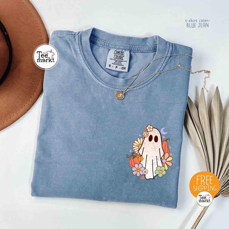 以下が含まれることがあります： 青いジーンズカラーのTシャツ。ゴーストのグラフィックが特徴です。ゴーストは花、カボチャ、三日月で飾られています。Tシャツは丸いネックラインと半袖です。「Tee markt」の文字が見えます。
