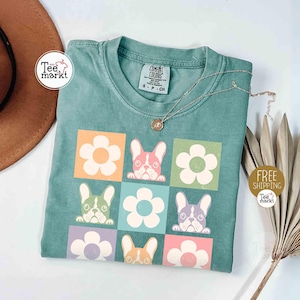 May include: A teal-colored t-shirt featuring a grid design with cartoon French bulldog faces and flower graphics in various colors. The shirt has a crew neck and short sleeves. A silver necklace with a pendant is draped across the shirt.